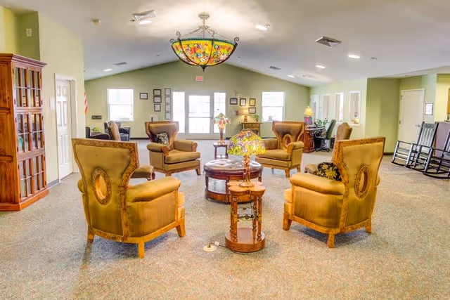 A spacious, well-lit common area with a high ceiling featuring a colorful stained glass chandelier. The room has several vintage-style armchairs arranged in a circle around a round wooden coffee table with a decorative lamp on it. There are windows letting in natural light, a wooden cabinet on the left, and rocking chairs along the right wall. The walls are painted light green and the carpet is a neutral tone.