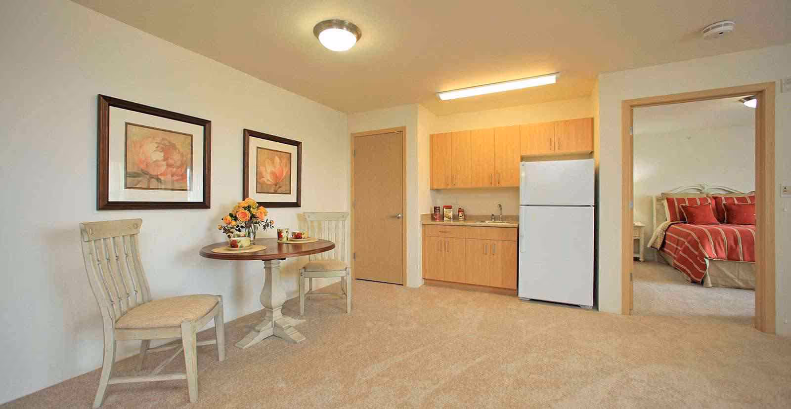 A small dining area with a round wooden table and two chairs, decorated with a vase of flowers and two place settings. To the right, there is a kitchenette with wooden cabinets, a sink, and a white refrigerator. An open doorway reveals a bedroom with a bed dressed in red and beige striped bedding. The room has beige carpeting and light-colored walls.
