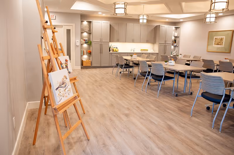 A spacious, well-lit room with wooden flooring featuring several tables and chairs arranged for group seating. On the left side, there are wooden easels displaying paintings of birds. The back wall has built-in cabinets and shelves with decorative items and plants. Ceiling lights provide warm illumination.