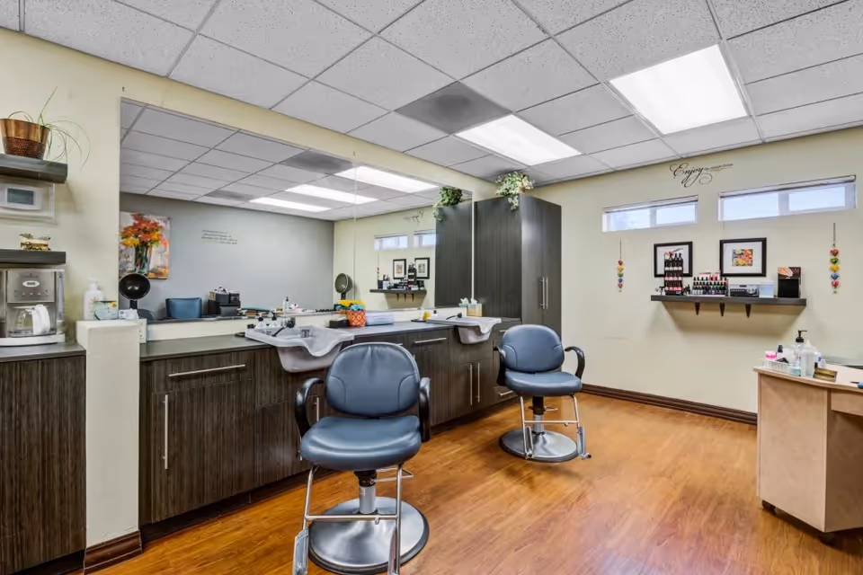 Interior view of a salon area in a senior living facility with two blue salon chairs in front of a long counter with sinks and large mirror. The room has wooden flooring, light-colored walls, and shelves with nail polish and other salon supplies. There are small windows near the ceiling and decorative plants on top of the cabinets.