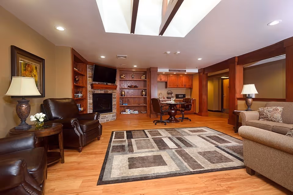A cozy living room area with wooden floors, a patterned area rug, two dark leather armchairs, a beige sofa with decorative pillows, and two table lamps on side tables. The room features a stone fireplace with a mounted TV above it, built-in wooden shelves, and a small dining table with chairs near a kitchenette with wooden cabinets. The ceiling has recessed lighting and a skylight.