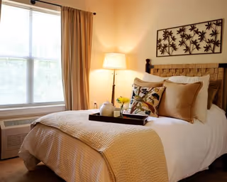 A cozy bedroom with a neatly made bed featuring beige and white bedding, decorative pillows, and a tray with a teapot and flowers. A floor lamp is lit beside the bed, and a large window with beige curtains lets in natural light. A decorative metal wall art piece hangs above the headboard.