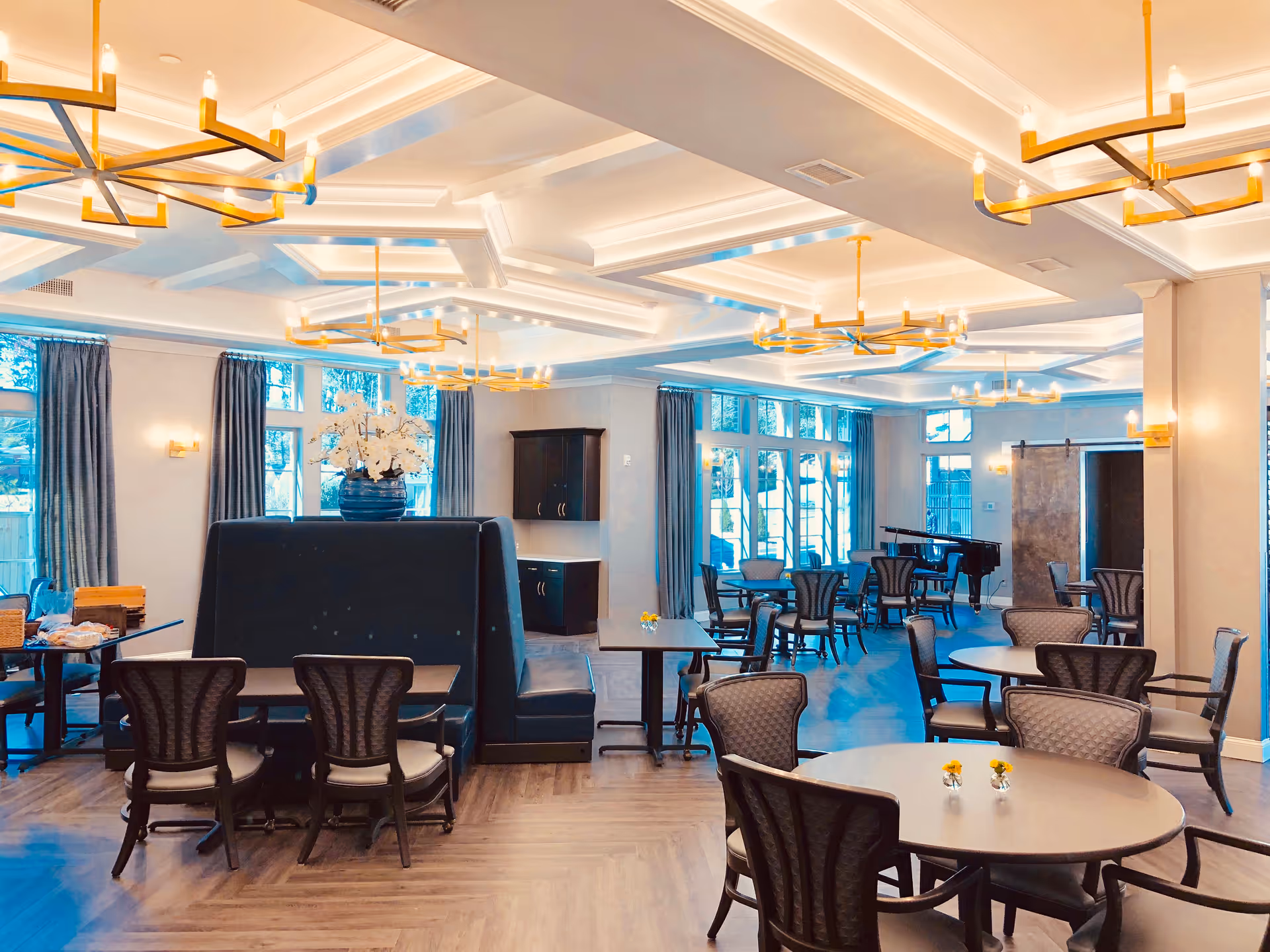 Spacious, well-lit dining and activity room with round tables and chairs, chandeliers, a piano, and large windows.