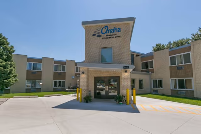Front exterior view of Omaha Nursing and Rehabilitation Center building with a clear blue sky, beige brick walls, and multiple windows. The entrance has double glass doors with a small covered area supported by columns and some plants near the entrance.