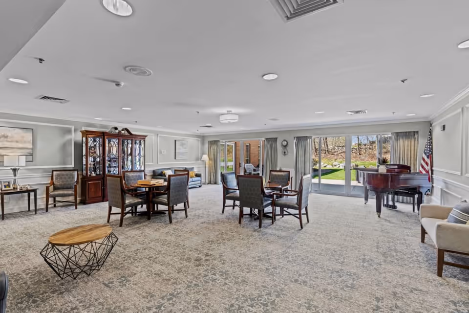 A spacious senior living common area with carpeted floor, several round tables with chairs, a wooden display cabinet, a sofa, armchairs, a grand piano, and large windows with curtains letting in natural light and showing an outdoor garden area.