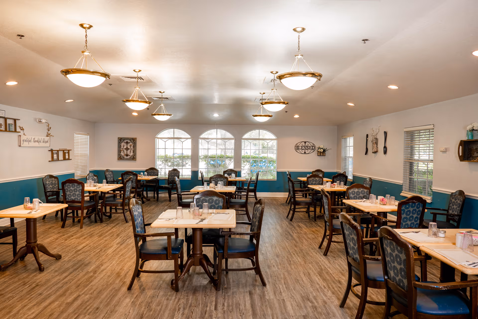 A dining room with multiple wooden tables and cushioned chairs arranged neatly. The room has large windows letting in natural light, teal and white walls, wooden flooring, and ceiling lights hanging above the tables. The tables are set with glasses, napkins, and condiments.