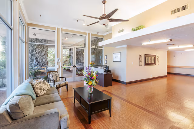 A spacious and bright living room area with large windows letting in natural light. The room features a gray sofa with cushions, a wooden coffee table with a vase of colorful flowers, and two striped armchairs. The floor is wooden, and there is a ceiling fan above. The background shows glass doors leading to an outdoor area with stone walls.