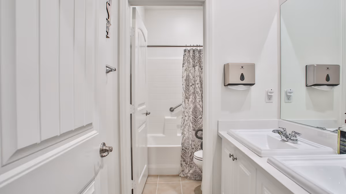Bright white bathroom with a double-sink vanity, wall-mounted soap dispensers, and a shower-tub with a patterned curtain and grab bar.