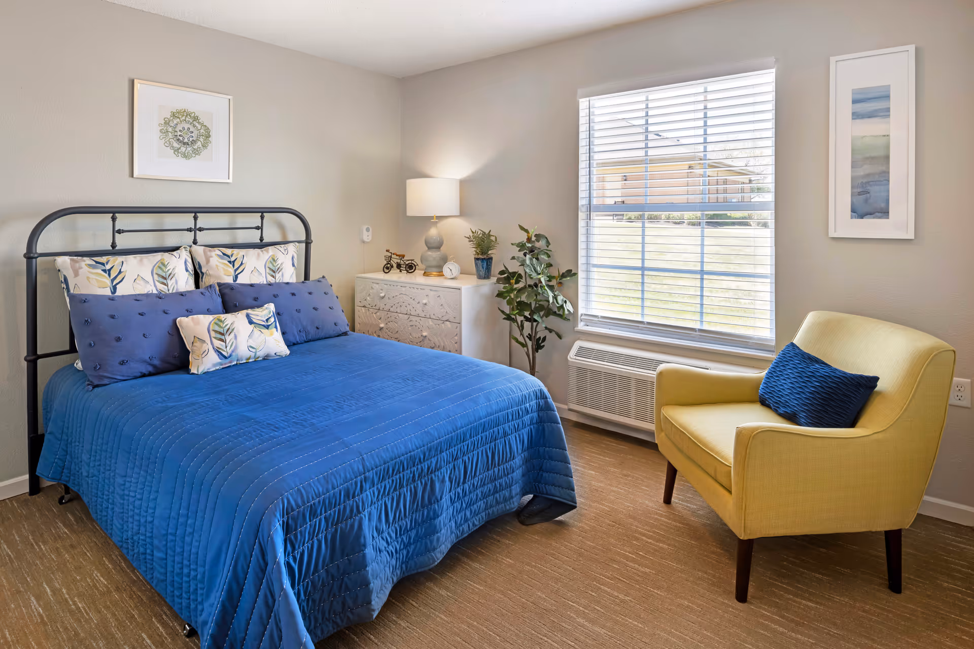 Bright furnished bedroom with a blue bedspread, metal bedframe, bedside dresser and a yellow armchair by a window.