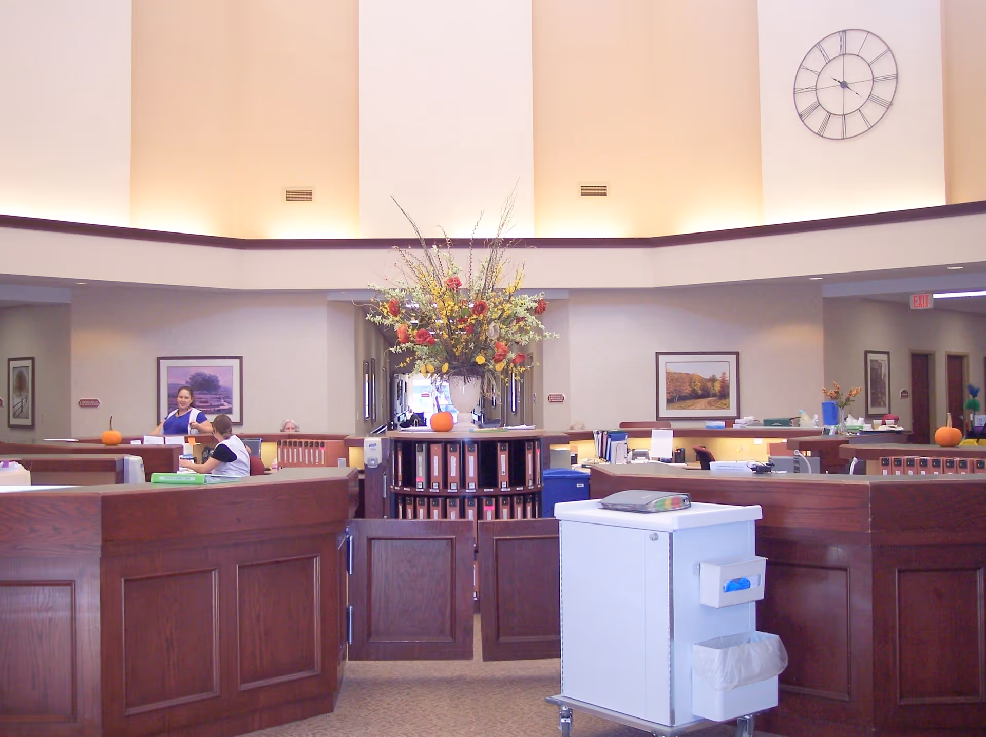 Reception area inside Lantern Park Specialty Care with a large flower arrangement on a central desk surrounded by wooden counters. Two staff members are visible behind the counters, and framed landscape pictures hang on the walls. A large clock is mounted high on the wall, and a white mobile cart is positioned in the foreground.