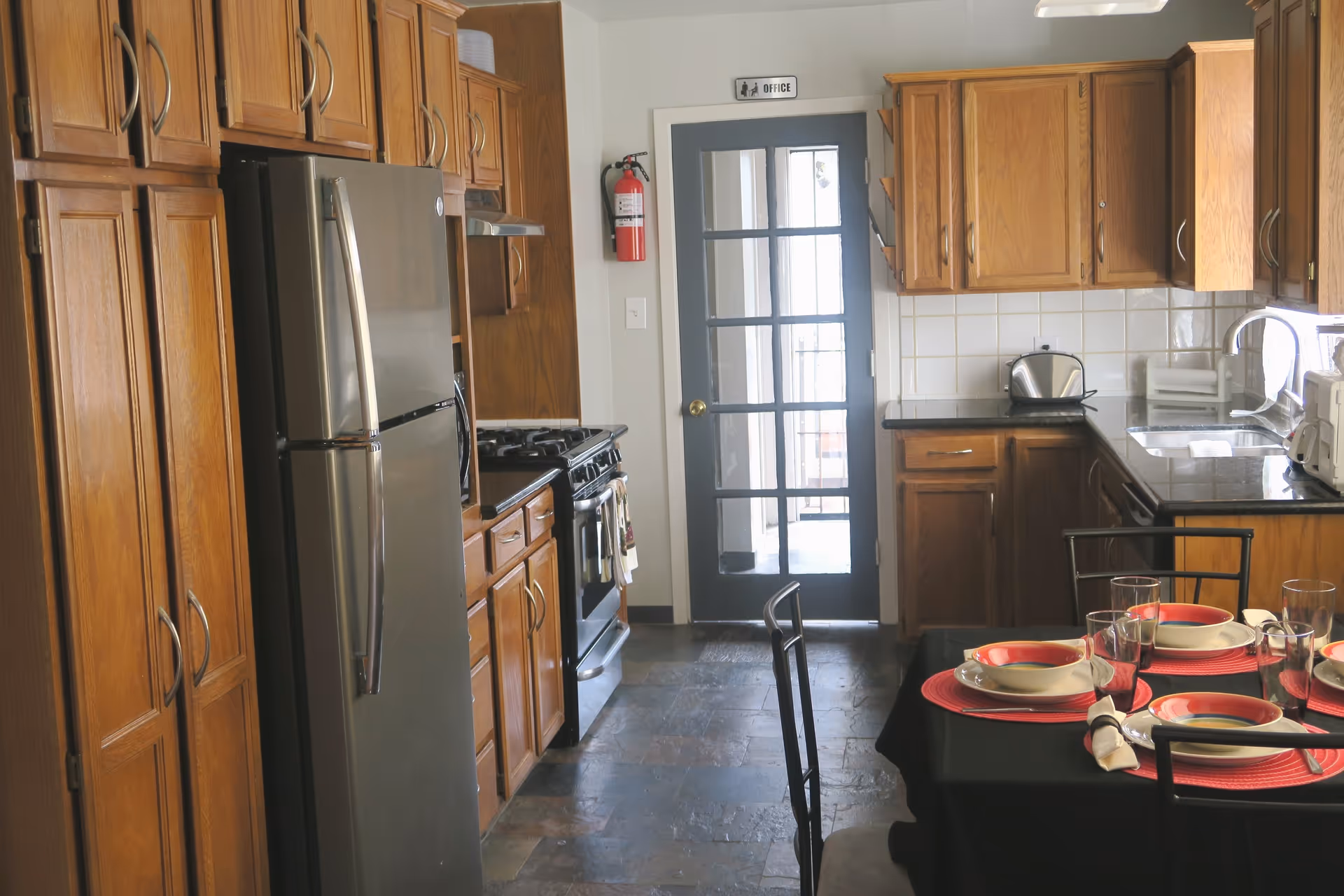 A kitchen with wooden cabinets, a stainless steel refrigerator, a stove, and a countertop with a toaster and other appliances. A dining table is set with red placemats, white bowls, plates, glasses, and napkins. A door with glass panels labeled 'Office' is visible in the background.
