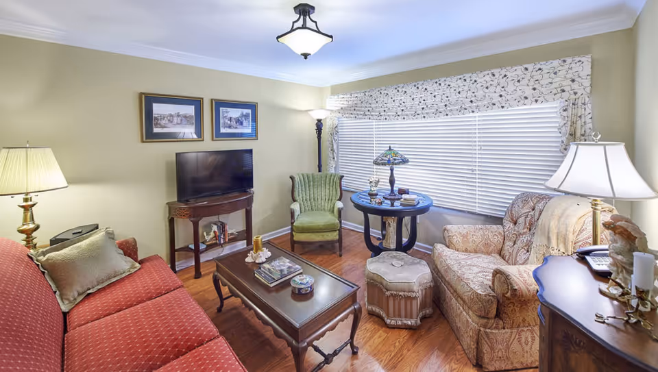 A cozy living room with a red sofa, a patterned armchair, a green upholstered chair, a wooden coffee table with books and decorative items, a round side table with a Tiffany-style lamp, and a TV on a wooden stand. The room has hardwood floors, beige walls, a large window with blinds and patterned valance, and multiple lamps providing warm lighting.