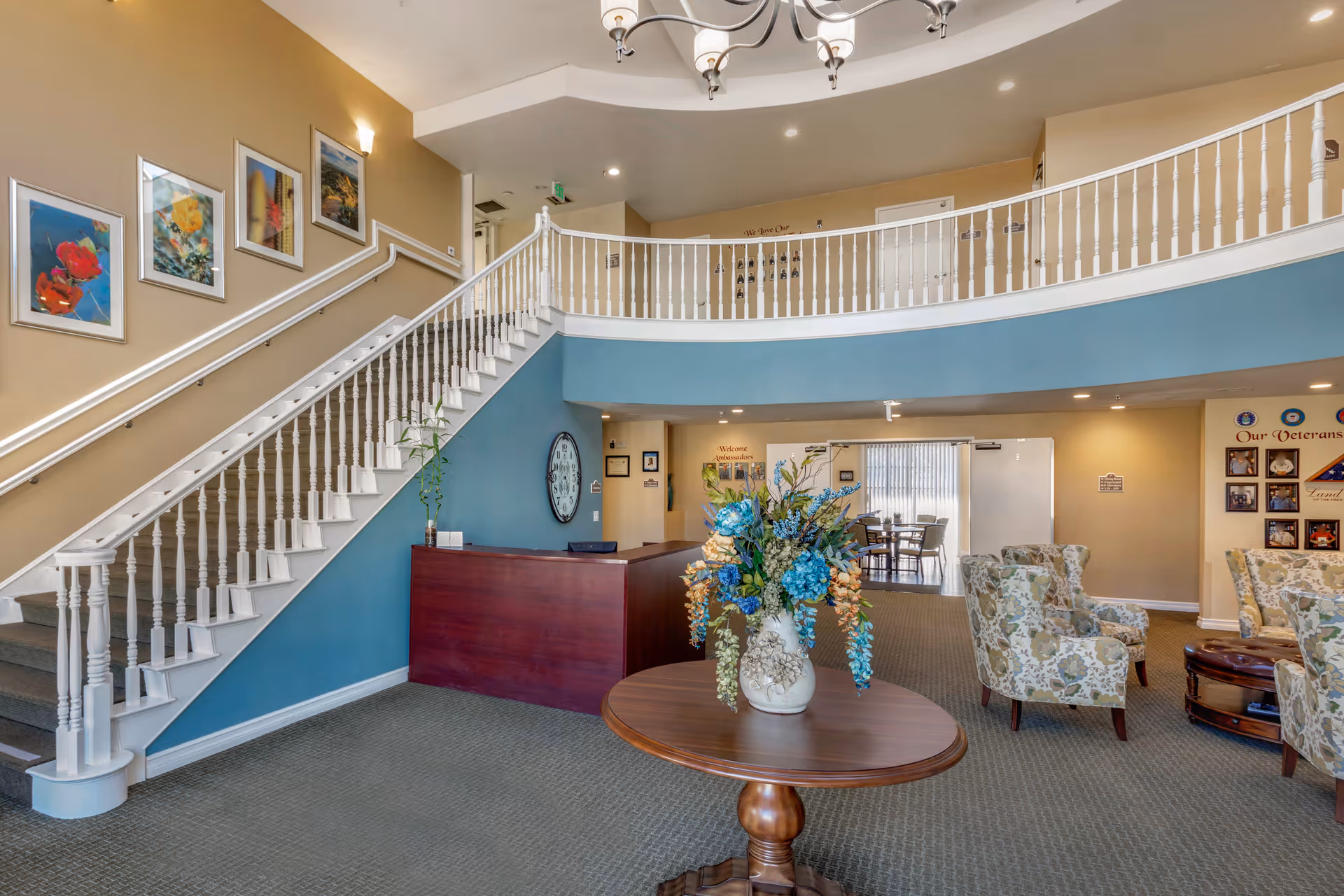 Two-story senior living lobby with a curved staircase and balcony, reception desk, seating area, and a central table holding a floral arrangement.