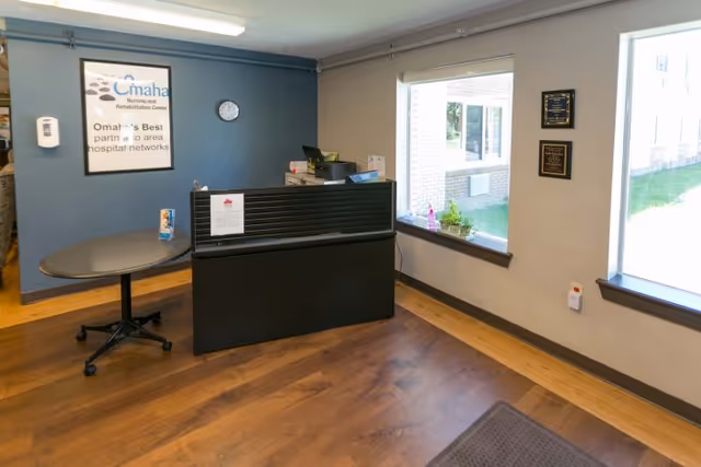 Reception area with a small black desk, a blue accent wall displaying an Omaha Nursing and Rehabilitation Center sign, and two windows.