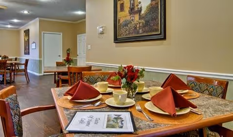 A dining area in a senior living facility with tables set for a meal. Each table has plates, cups, silverware, and red folded napkins. There is a small vase with red flowers on the table in the foreground, along with a menu. The room has beige walls, wooden chairs, and framed artwork on the walls.