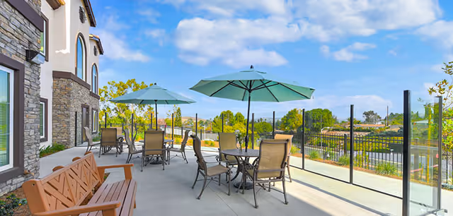 Outdoor patio area at Silverado Thousand Oaks featuring several tables with chairs and large green umbrellas. The patio is adjacent to a building with stone and stucco exterior walls. There is a wooden bench on the left side and glass panel fencing along the edge of the patio, overlooking greenery and trees under a partly cloudy blue sky.