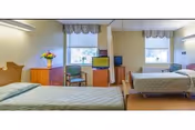 Shared senior living bedroom with two twin beds, wooden dressers, chairs, a TV, and two windows with valances.
