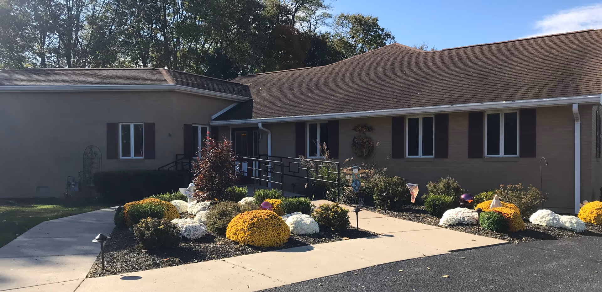 Exterior view of a single-story senior living facility building with a sloped roof, beige walls, and several windows with dark shutters. A concrete walkway leads to the entrance, which has a black metal handrail and a ramp. The landscaped garden in front features neatly arranged bushes and colorful flowers, including yellow and white blooms. Trees are visible in the background under a clear blue sky.