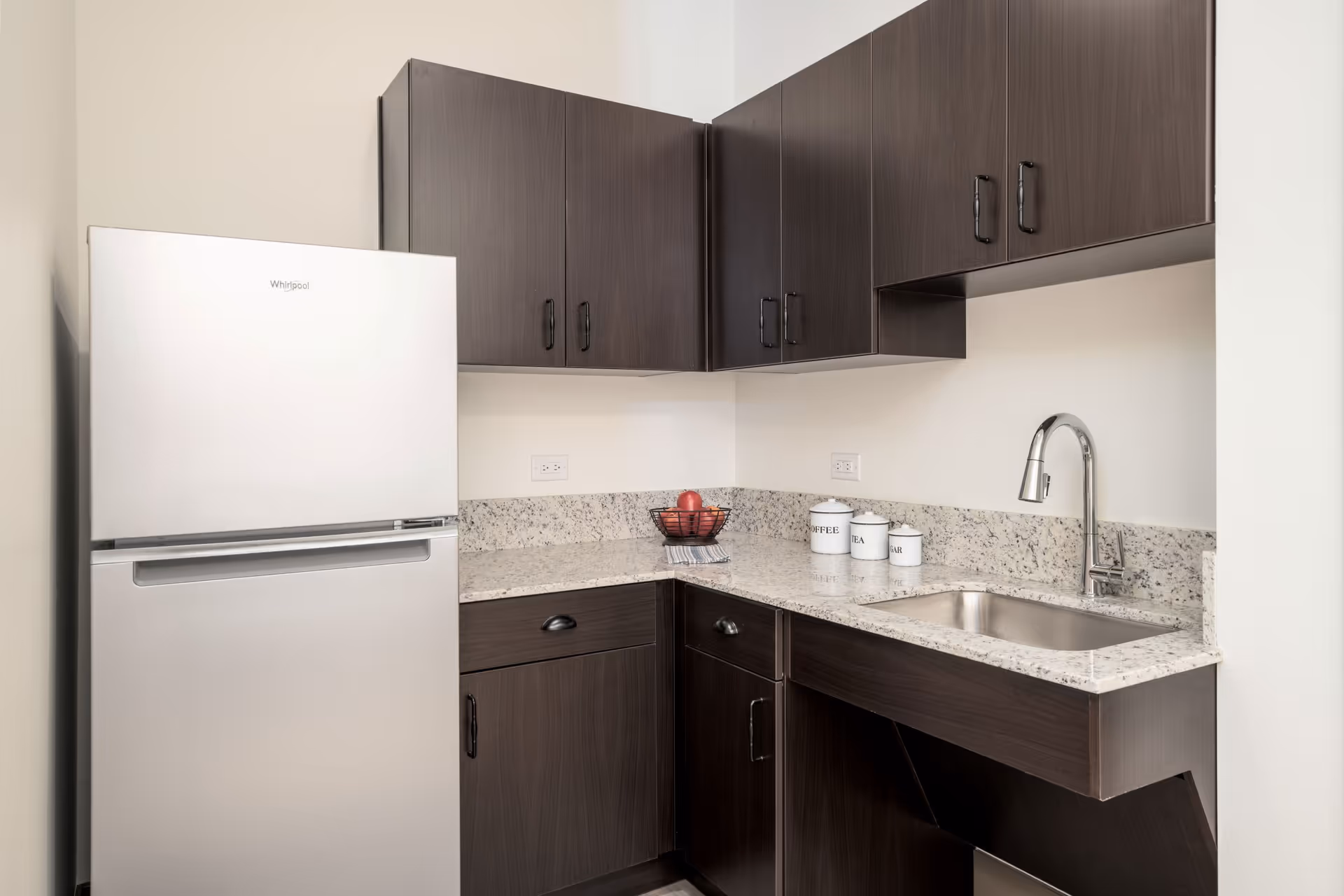 A small kitchen area with dark wood cabinets, a white Whirlpool refrigerator, a granite countertop with a stainless steel sink and faucet, and three white canisters labeled Coffee, Tea, and Sugar. There is also a wire basket with red apples on the counter.