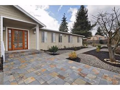 Exterior view of a single-story building with beige siding and a stone-tiled patio leading to a wooden door with glass panels. The surrounding area includes landscaped garden beds with small shrubs and trees under a partly cloudy sky.