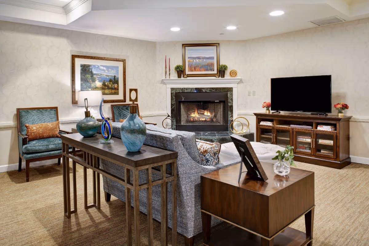 A cozy living room with a lit fireplace in the center, a gray sofa facing the fireplace, two teal armchairs with orange cushions, a wooden TV stand with a flat-screen TV, decorative vases and artwork on the walls, and a wooden side table with a small plant and picture frame.