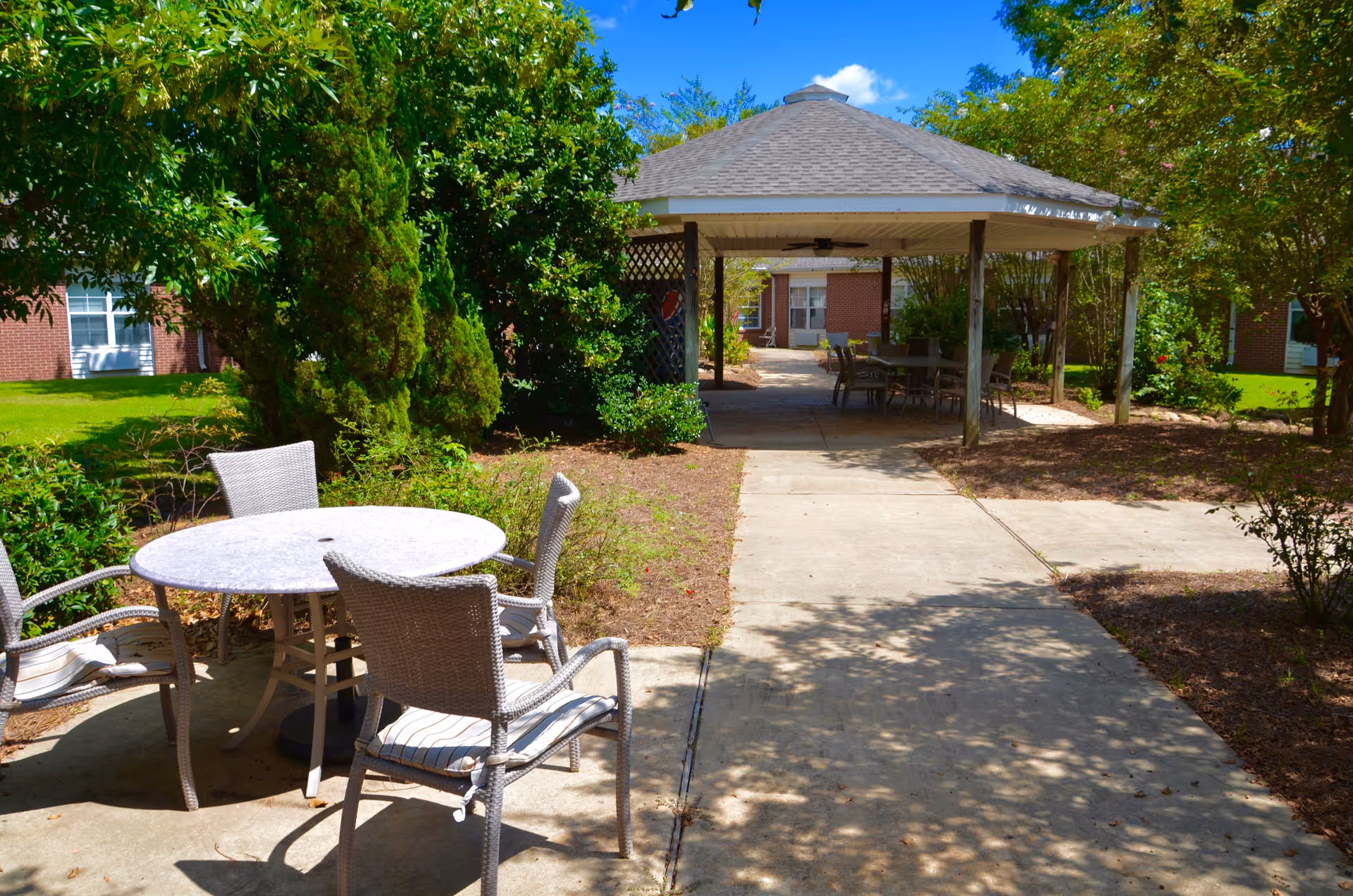 Sunlit courtyard with a round patio table and chairs in the foreground and a covered gazebo with additional seating at the end of a concrete path.