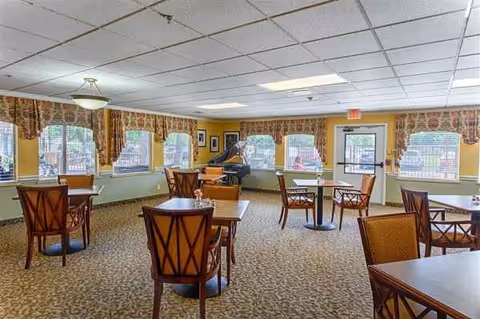 Empty dining room with tables, chairs, windows, and a piano in a senior living facility.