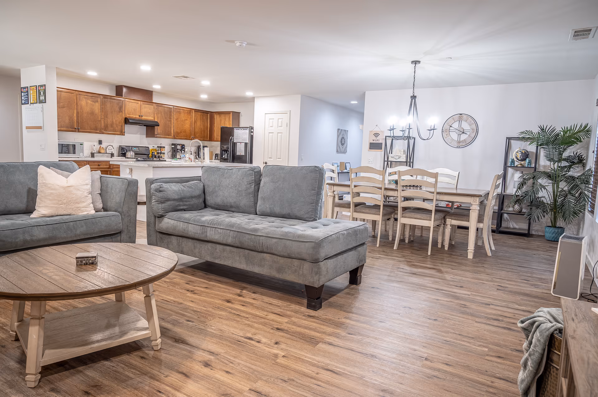 A bright and spacious open-concept living area featuring two gray sofas with cushions, a round wooden coffee table, a dining table with six chairs, a modern chandelier, a large wall clock, and a kitchen with wooden cabinets and stainless steel appliances in the background. The room has wooden flooring and decorative plants.