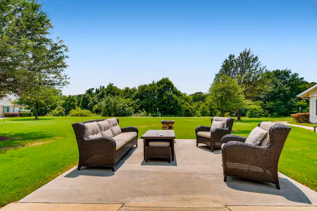 Outdoor patio area with a set of brown wicker furniture including a sofa, two armchairs, a coffee table, and a small side table, all with beige cushions, set on a concrete slab surrounded by green grass and trees under a clear blue sky.