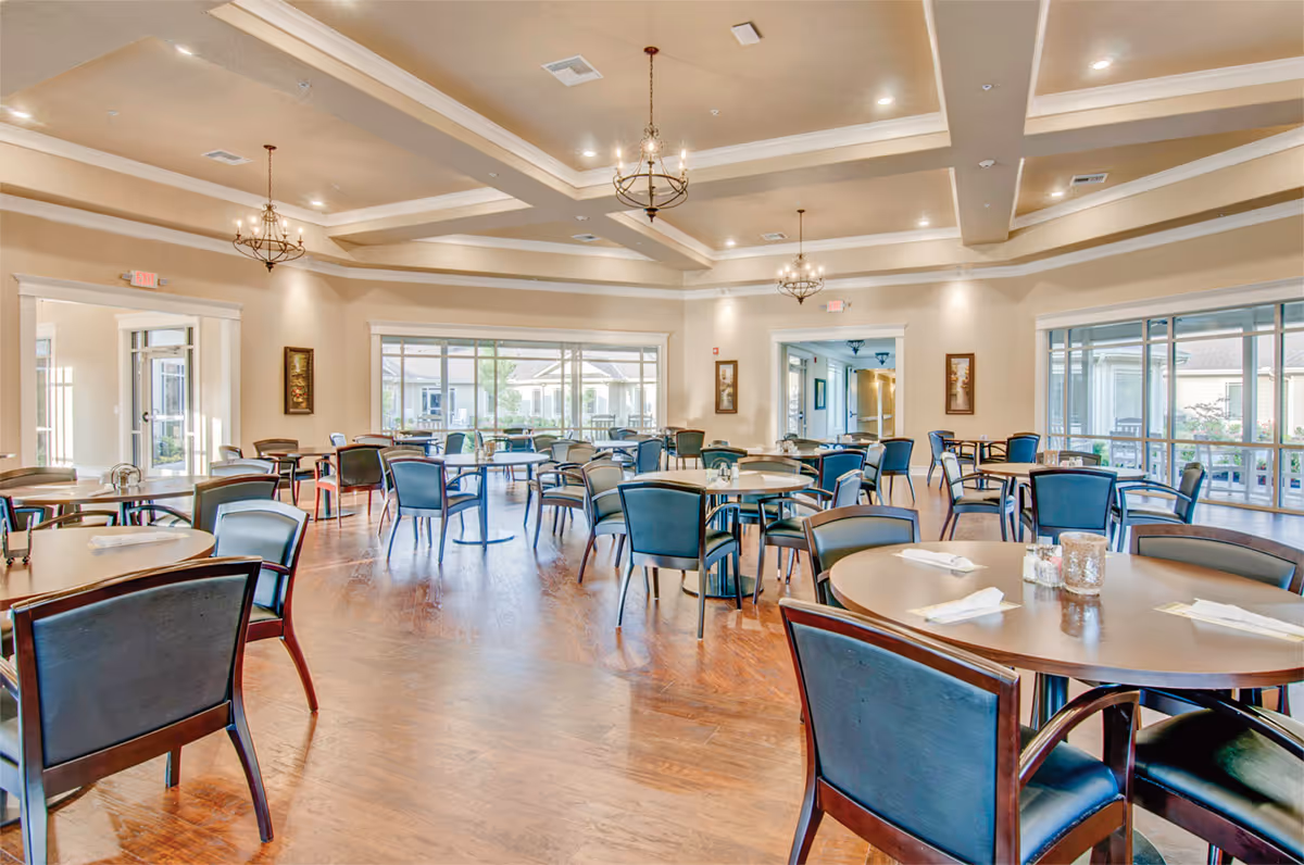 A spacious dining room with multiple round and square tables surrounded by dark wooden chairs with black cushions. The room features large windows allowing natural light to fill the space, beige walls, and a coffered ceiling with elegant chandeliers. The floor is made of polished wood, and the room appears clean and well-organized.