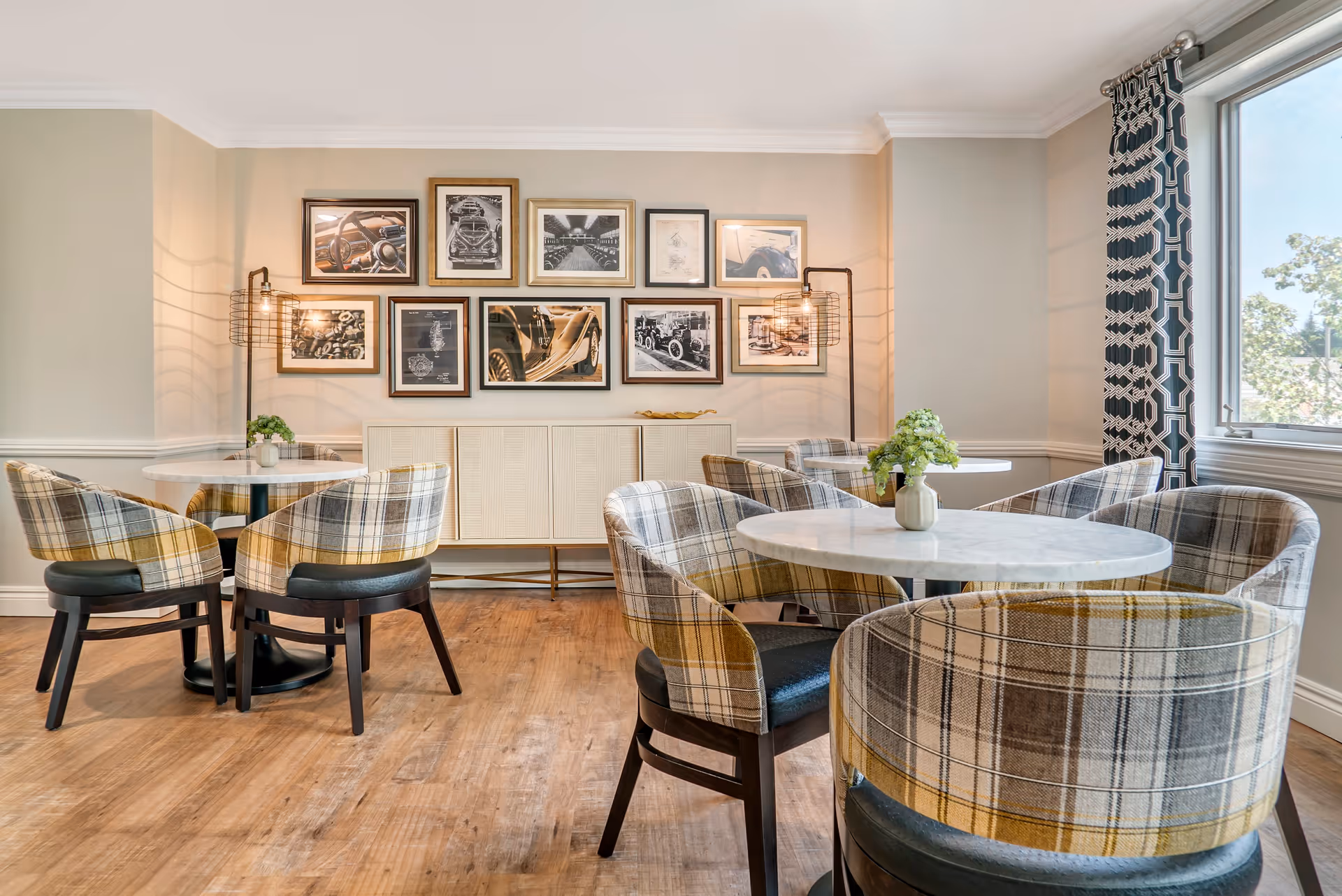 A cozy dining area with round white marble tables and plaid upholstered chairs. The room features wooden flooring, a large window with patterned curtains, and a wall decorated with framed black and white photographs. Two modern wall lamps with exposed bulbs provide warm lighting.