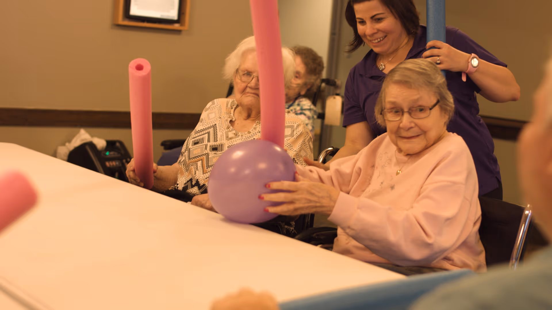 Two elderly women sitting at a table holding pink pool noodles and a purple ball, with a caregiver standing behind them smiling and holding a blue pool noodle in a warm indoor setting.