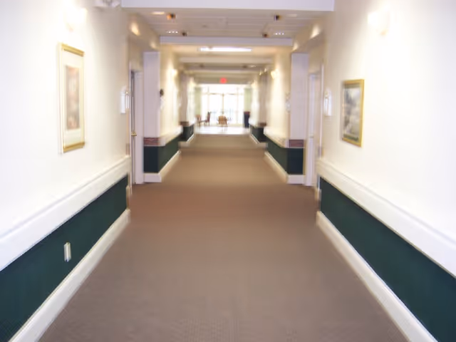 A long, well-lit hallway in a facility with beige carpet, white walls with a dark green lower section, framed pictures on the walls, and doors on both sides leading to rooms. At the end of the hallway, there is a seating area with chairs near large windows letting in natural light.