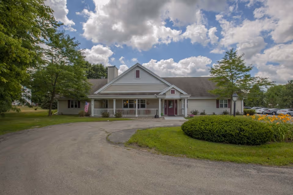 Single-story assisted living building with a covered front porch, circular driveway, landscaped lawn and trees under a cloudy sky.