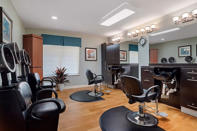 A well-lit salon area with multiple black salon chairs and hair washing stations. The room has wooden flooring, light-colored walls, a large mirror, and cabinets for storage. There are framed pictures on the walls and a potted plant near a window with a blue valance.