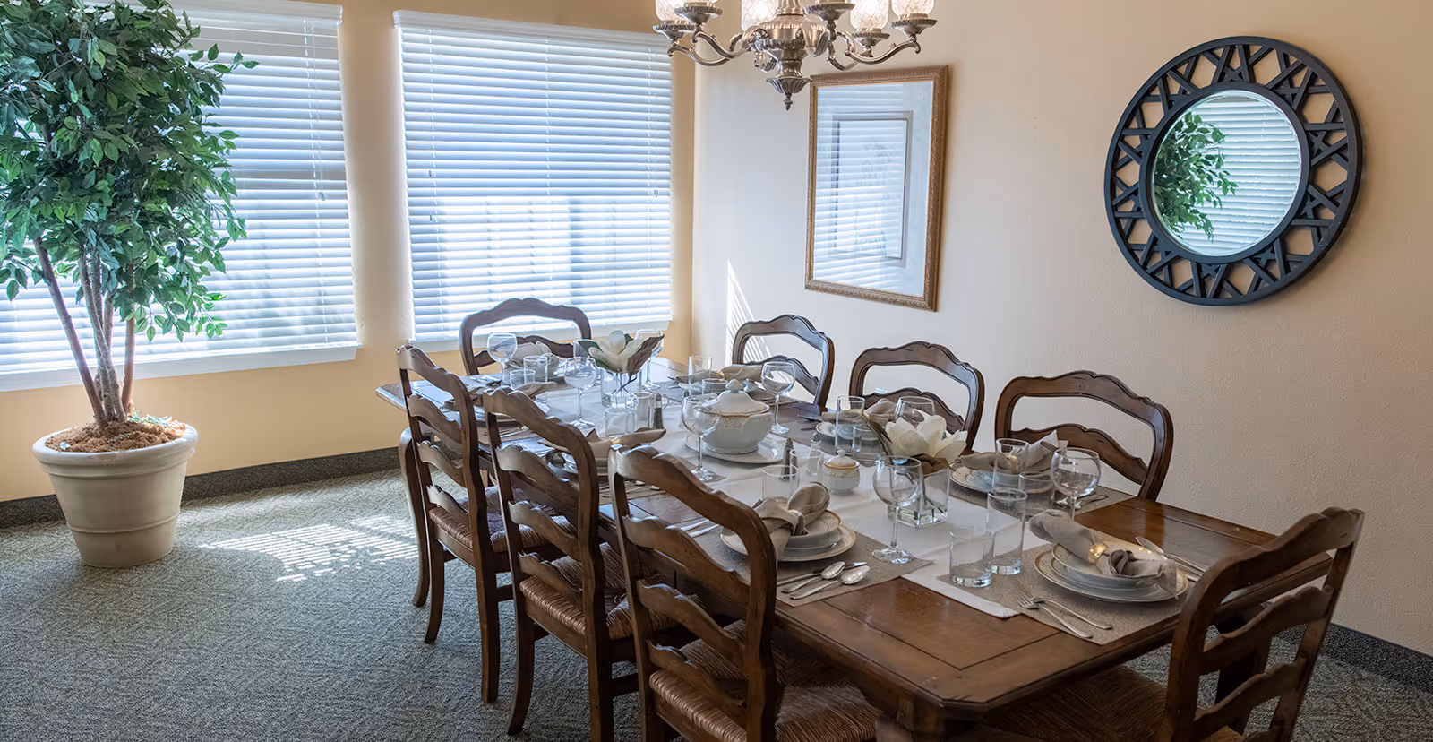 Sunlit dining room with a long wooden table set for eight, wooden chairs, a potted plant, and a decorative wall mirror.
