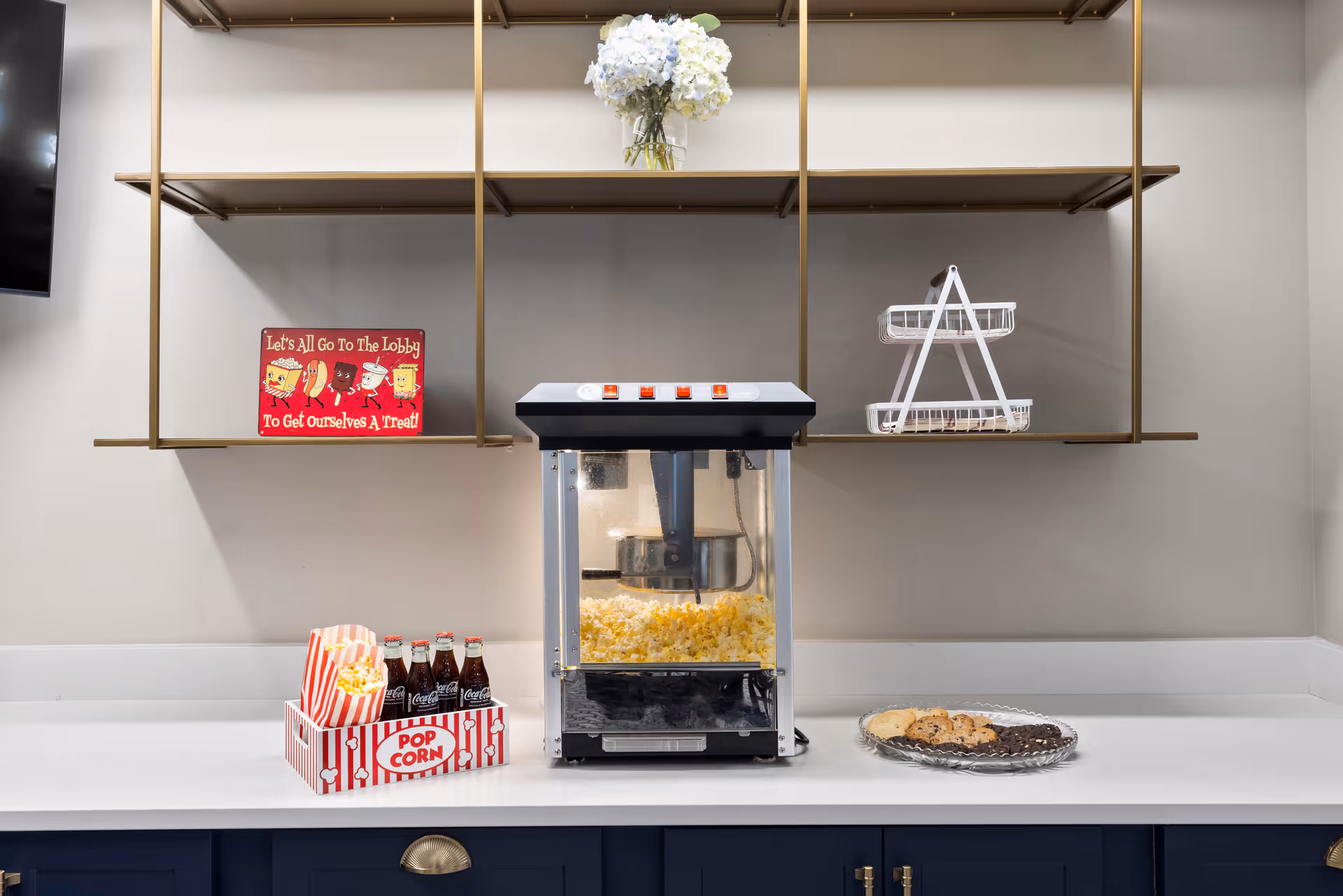 A countertop with a popcorn machine filled with popcorn, a tray holding popcorn bags and bottles of Coca-Cola, and a plate of assorted cookies. Above the counter are gold-colored shelves, one holding a vase with white flowers and another with a small red sign that reads 'Let's All Go To The Lobby To Get Ourselves A Treat'.