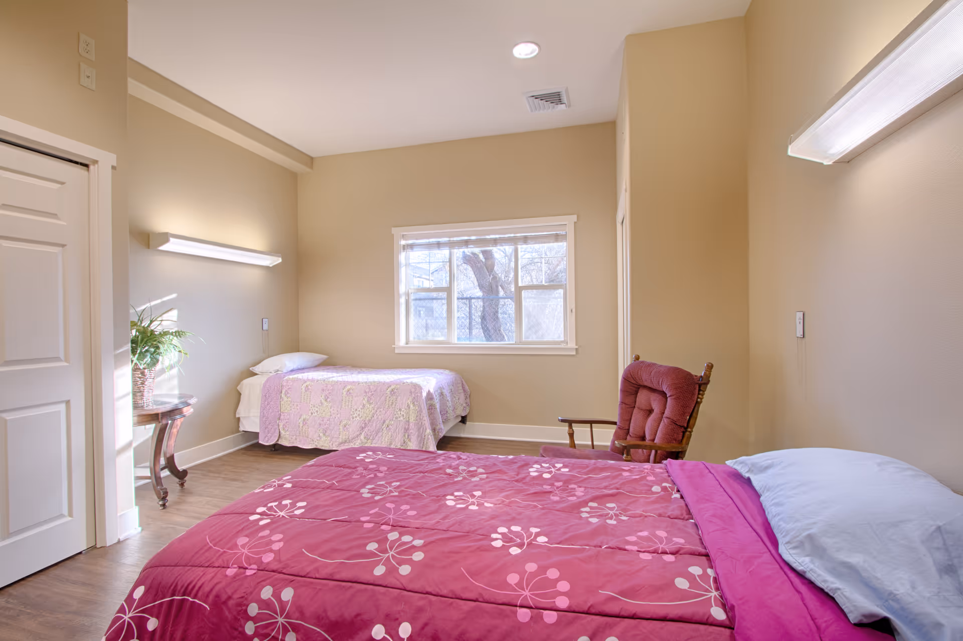 A bright bedroom with two single beds, one with a pink floral bedspread and the other with a light purple quilt. There is a wooden rocking chair with a red cushion between the beds, a small round table with a potted plant near the window, and beige walls with white trim. A window lets in natural light, and the floor is wood.