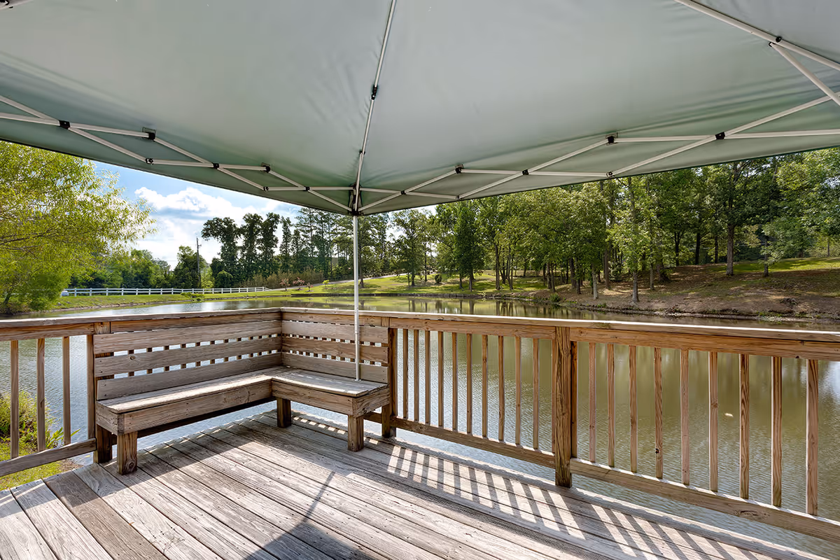 A wooden deck with built-in bench seating under a large canopy, overlooking a calm pond surrounded by trees and greenery on a sunny day.