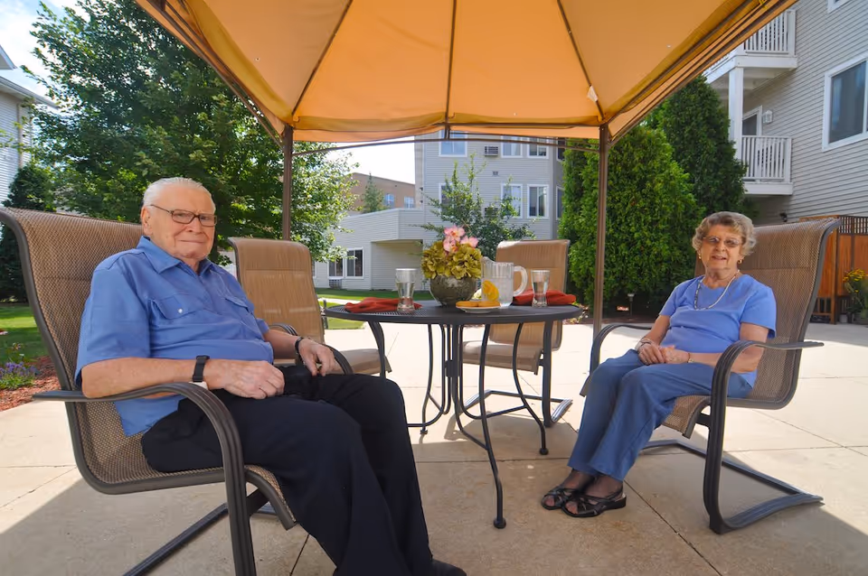 An elderly man and woman sitting on patio chairs under a canopy at a round table with a flower pot, pitcher of water with lemon slices, and glasses, outside a residential building with trees and shrubs in the background.