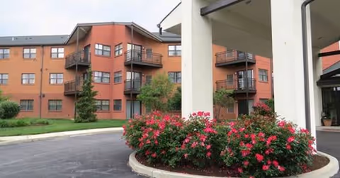 Red brick senior living building with balconies, curved driveway, and blooming shrubs.