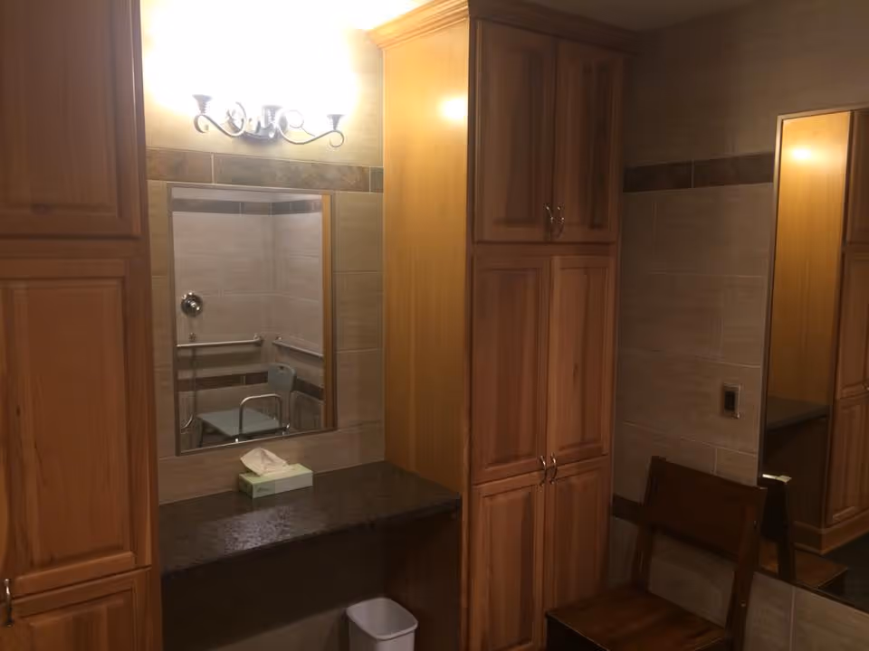 Interior view of a bathroom area with wooden cabinets, a granite countertop with a tissue box, a wall-mounted mirror reflecting a shower area with a shower chair and grab bars, a wooden chair, and a wall-mounted light fixture above the mirror.