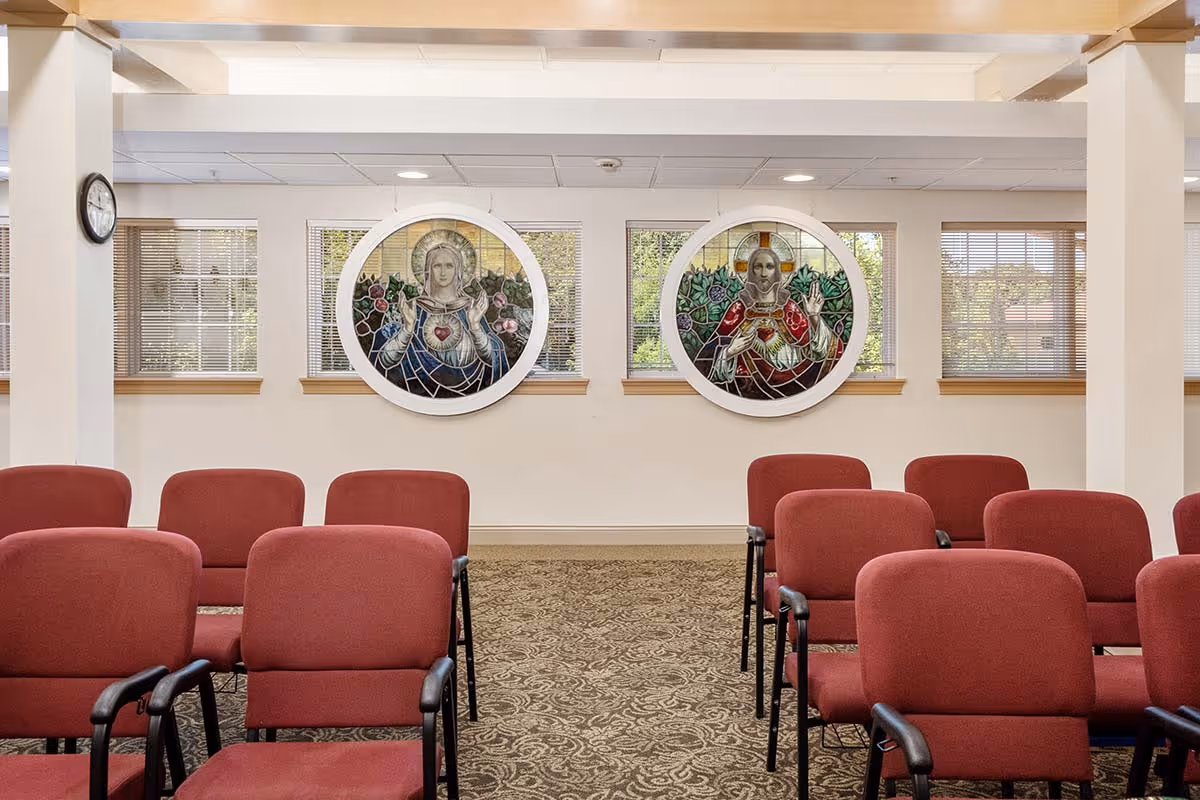 Interior room with rows of red cushioned chairs arranged facing a wall with two round stained glass windows depicting religious figures. The room has beige walls, windows with blinds, and a patterned carpet.