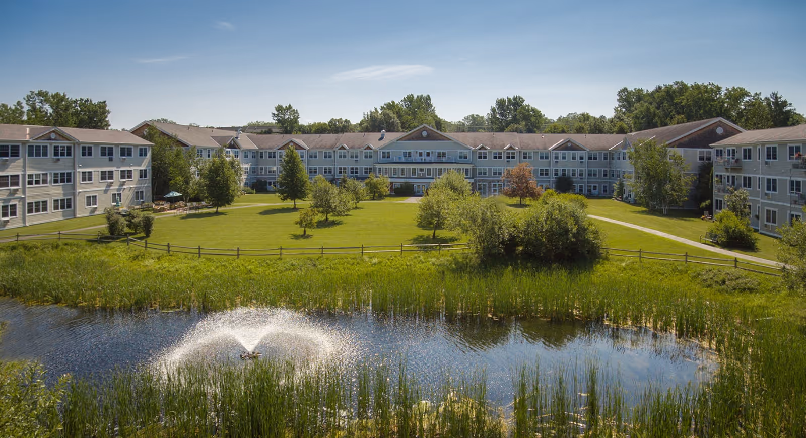 A large senior living facility building with three stories, surrounded by green lawns, trees, and a small pond with a water fountain in the foreground under a clear blue sky.
