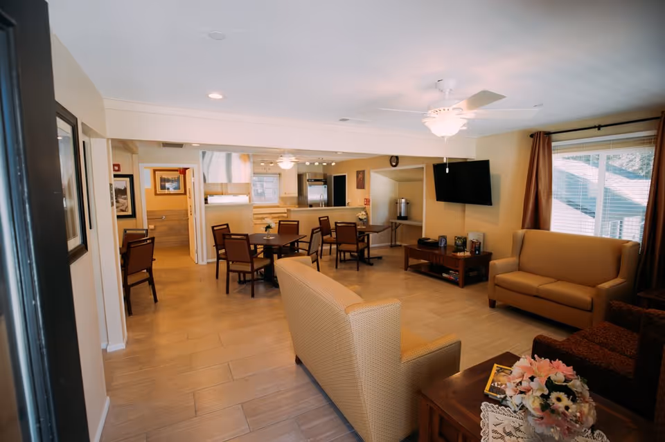 A bright and spacious common area in a senior living facility featuring a seating area with sofas and armchairs, a coffee table with a flower arrangement, a wall-mounted TV, and a dining area with tables and chairs near a kitchen with stainless steel appliances.