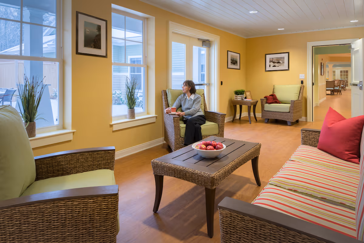 A cozy living room area in a senior living facility with wicker furniture including a striped sofa with red pillows, two armchairs with green cushions, and a wooden coffee table with a bowl of apples. A woman is sitting on one of the armchairs holding a cup and looking out the window. The walls are painted yellow and decorated with framed pictures. There are large windows letting in natural light and a door leading outside.