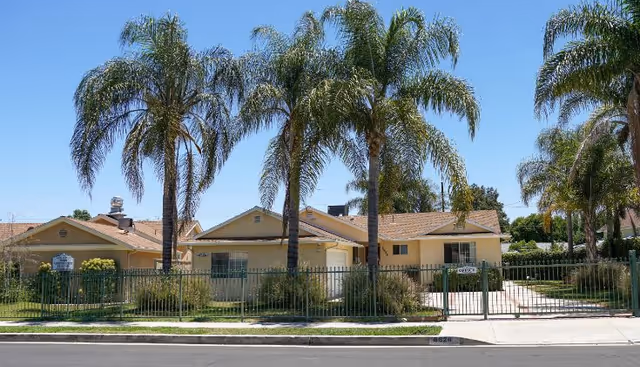 Single-story beige senior living building behind a metal fence with tall palm trees.