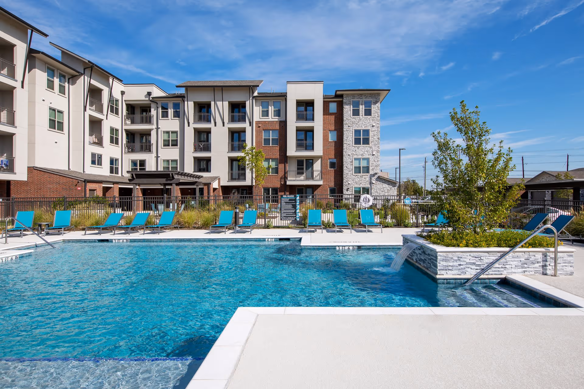 Outdoor swimming pool with clear blue water surrounded by turquoise lounge chairs. A modern multi-story building with balconies and a mix of brick and stone exterior is in the background under a blue sky with some clouds. There is a small raised planter with greenery and a water feature flowing into the pool.