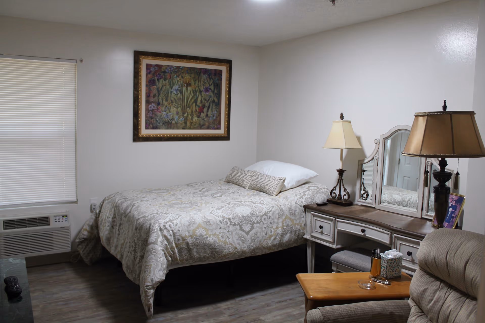 A cozy bedroom with a bed covered in a patterned beige and white comforter, a decorative pillow, and a white pillow. There is a framed floral painting on the wall above the bed. Next to the bed is a vintage-style dresser with a tri-fold mirror, two table lamps, and some small items including a tissue box and a glass. A comfortable armchair and a small wooden table are also visible. The room has a window with closed blinds and a wall-mounted air conditioning unit below it.