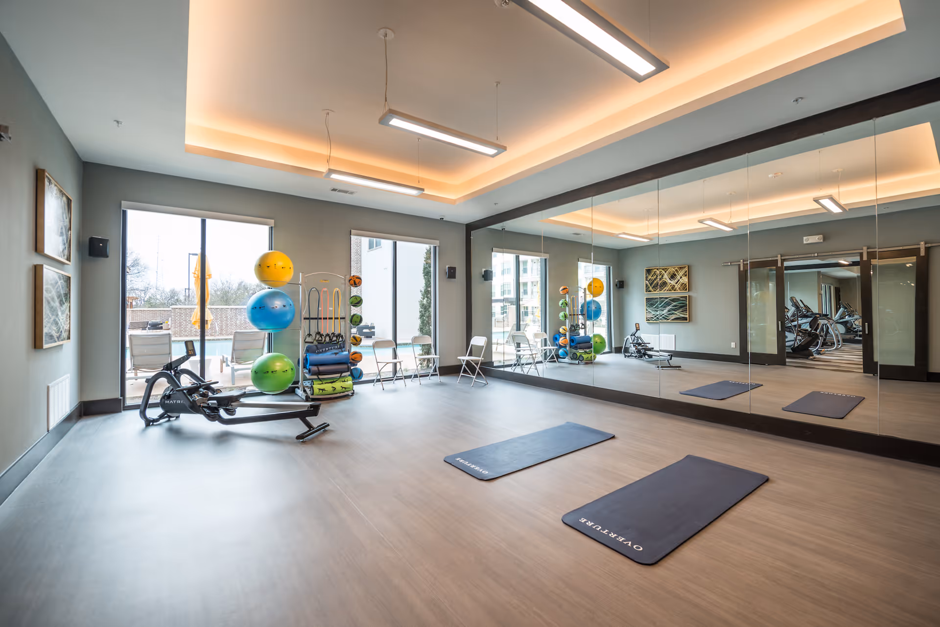 A bright exercise room with large mirrors on one wall, two exercise mats on the floor labeled 'OVERTURE', a rowing machine, and a rack holding colorful exercise balls, resistance bands, and other fitness equipment. The room has large windows with a view of an outdoor pool area and modern ceiling lights.